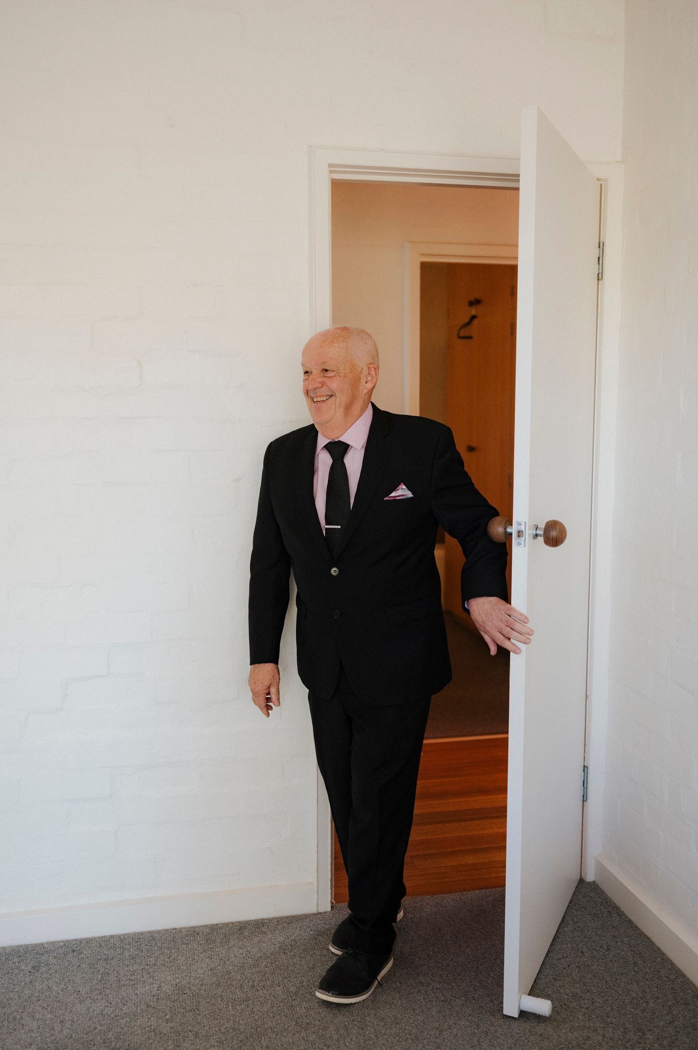 Elderly man in black suit smiling while opening a white door in a bright room.