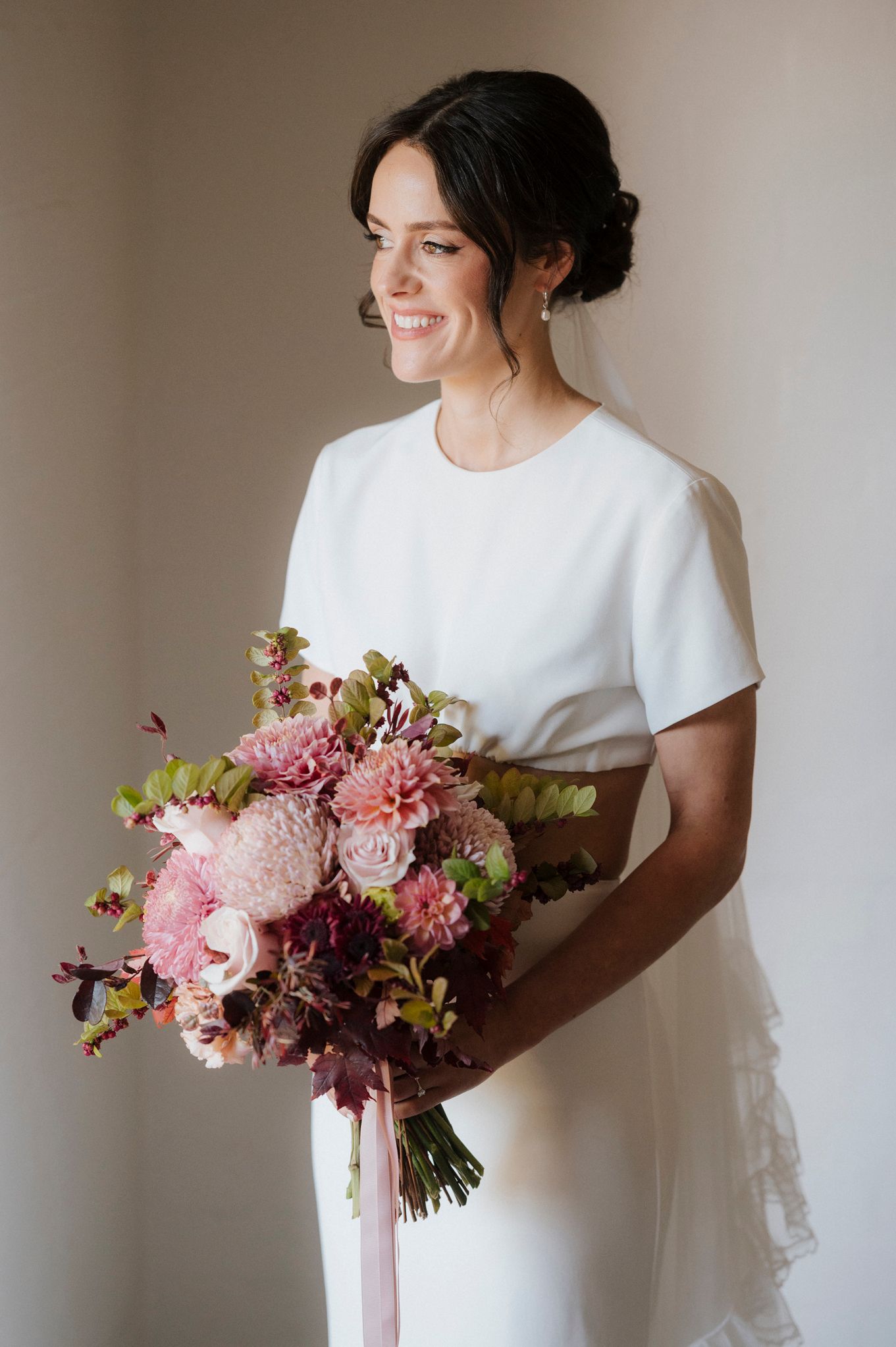 Smiling bride in white dress holding a lush pink and burgundy wedding bouquet with dahlias and roses.