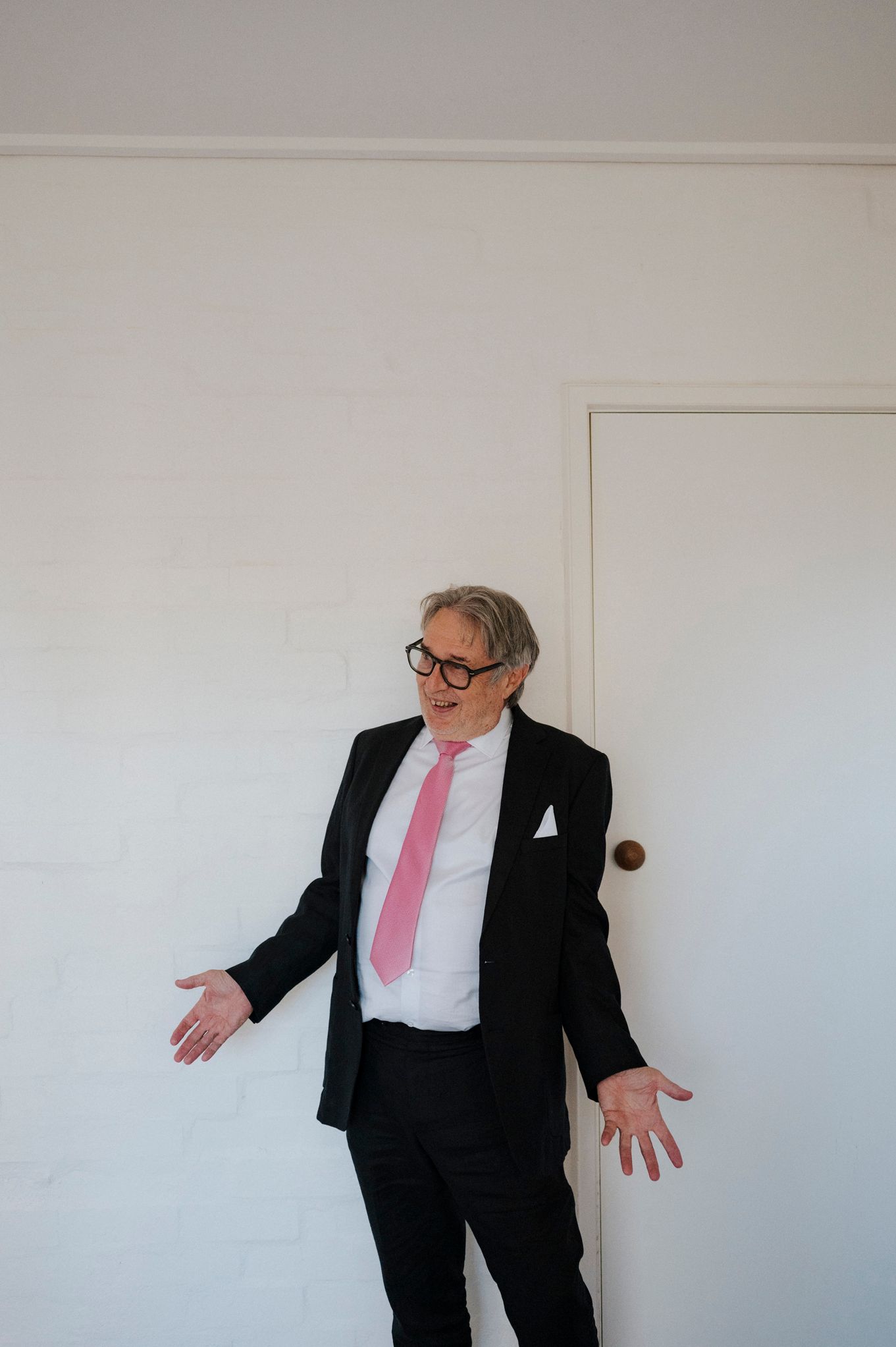 Older man in black suit and pink tie gesturing with open hands, wearing glasses, standing near a white door.