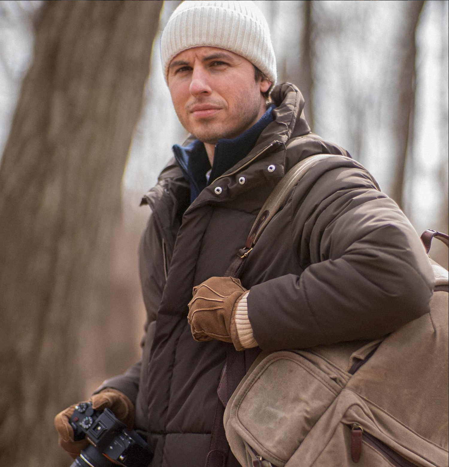Man in white beanie hat and brown jacket holding camera with leather gloves outdoors in winter.