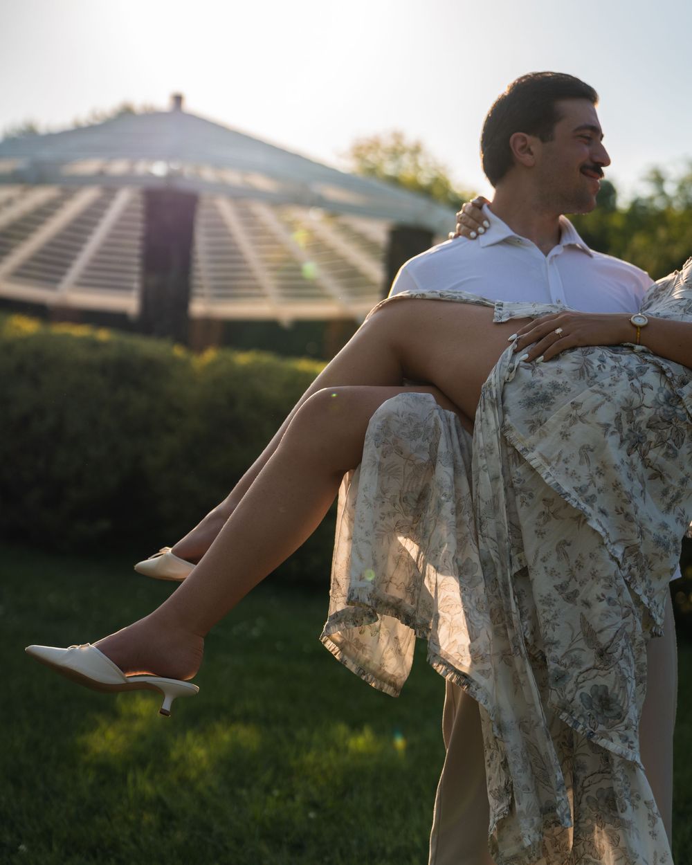 A groom carries his bride in a floral dress and white heels outdoors near a greenhouse in golden hour light.