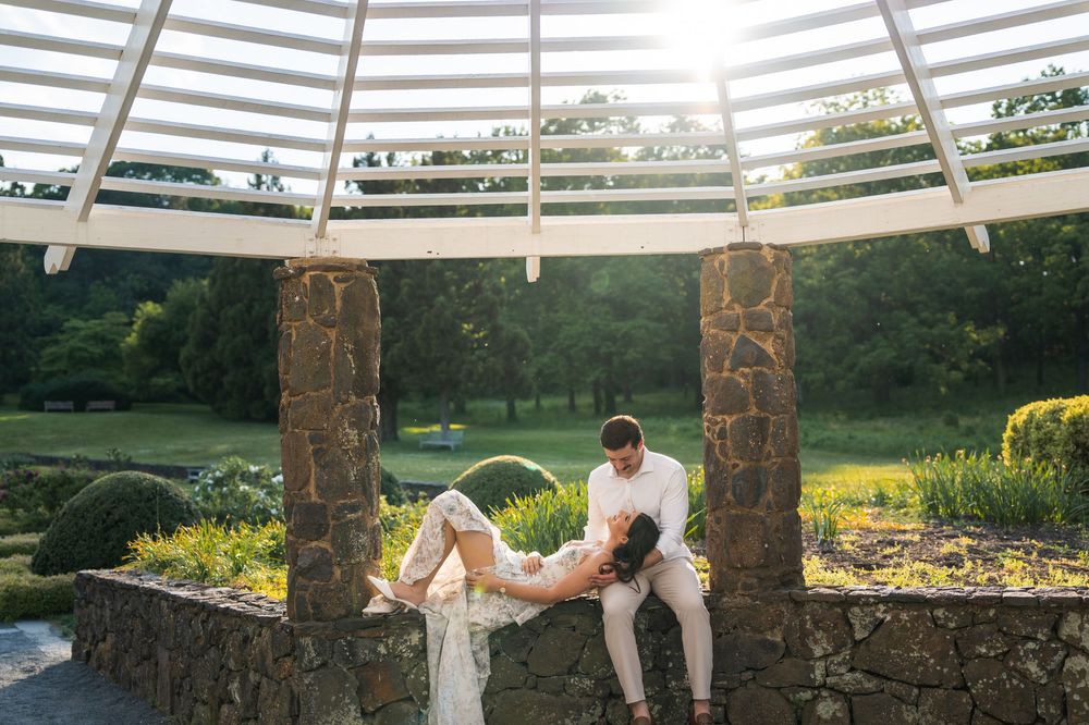 Bride and groom sharing a romantic moment under a stone pergola surrounded by lush gardens at golden hour.