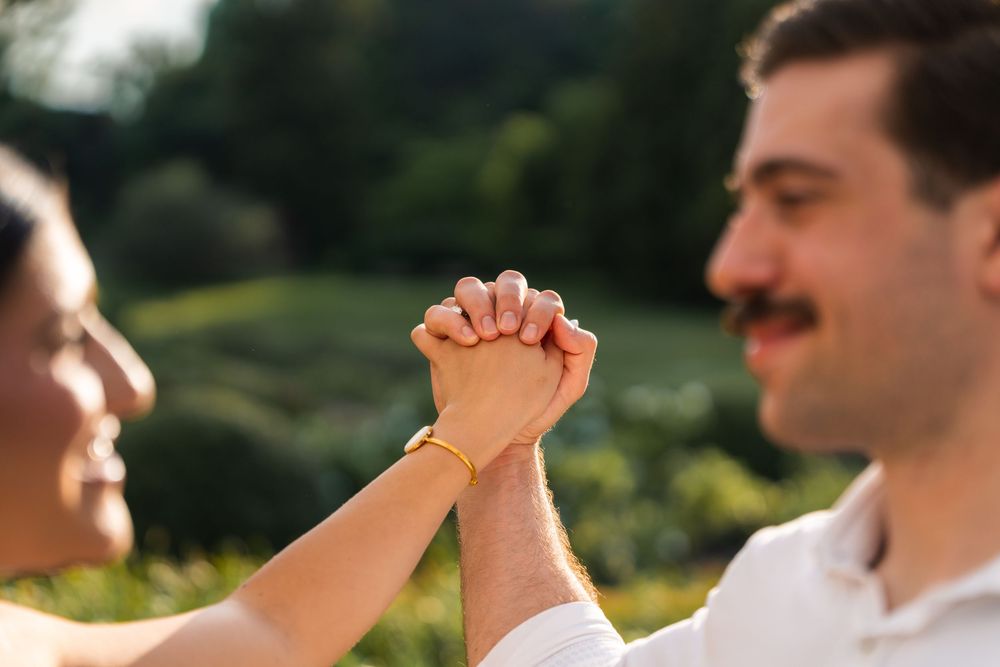 Couple holding hands outdoors in a sunny green park, man with mustache smiling, woman wearing gold bracelet.