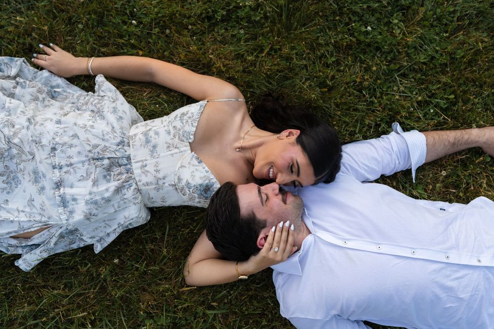 Couple lying on green grass, woman in floral dress leaning close to man in white shirt, sharing a tender moment.