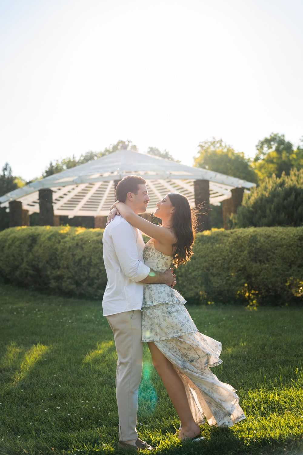 Couple embracing in a sunlit garden with a white pergola and lush green hedges in the background.