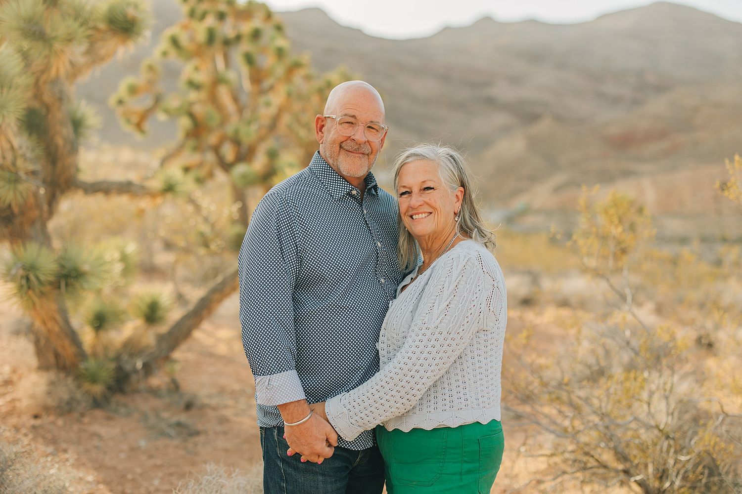 A smiling older couple holds hands in a desert landscape with Joshua trees and mountains in the background.