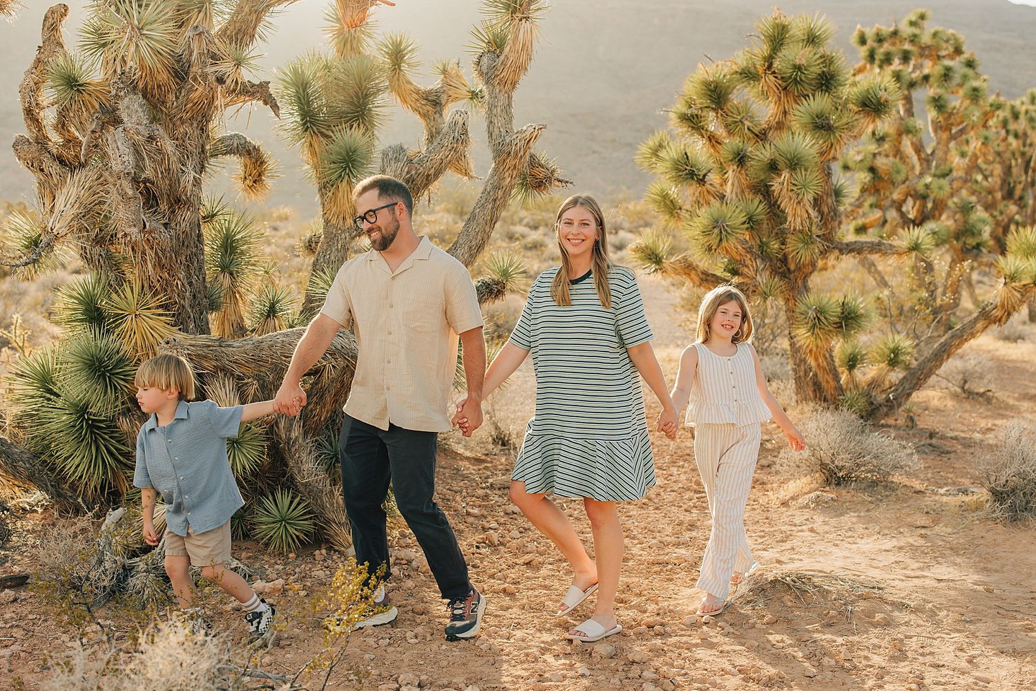 Family of four holding hands walking through Joshua Tree desert landscape with cacti.