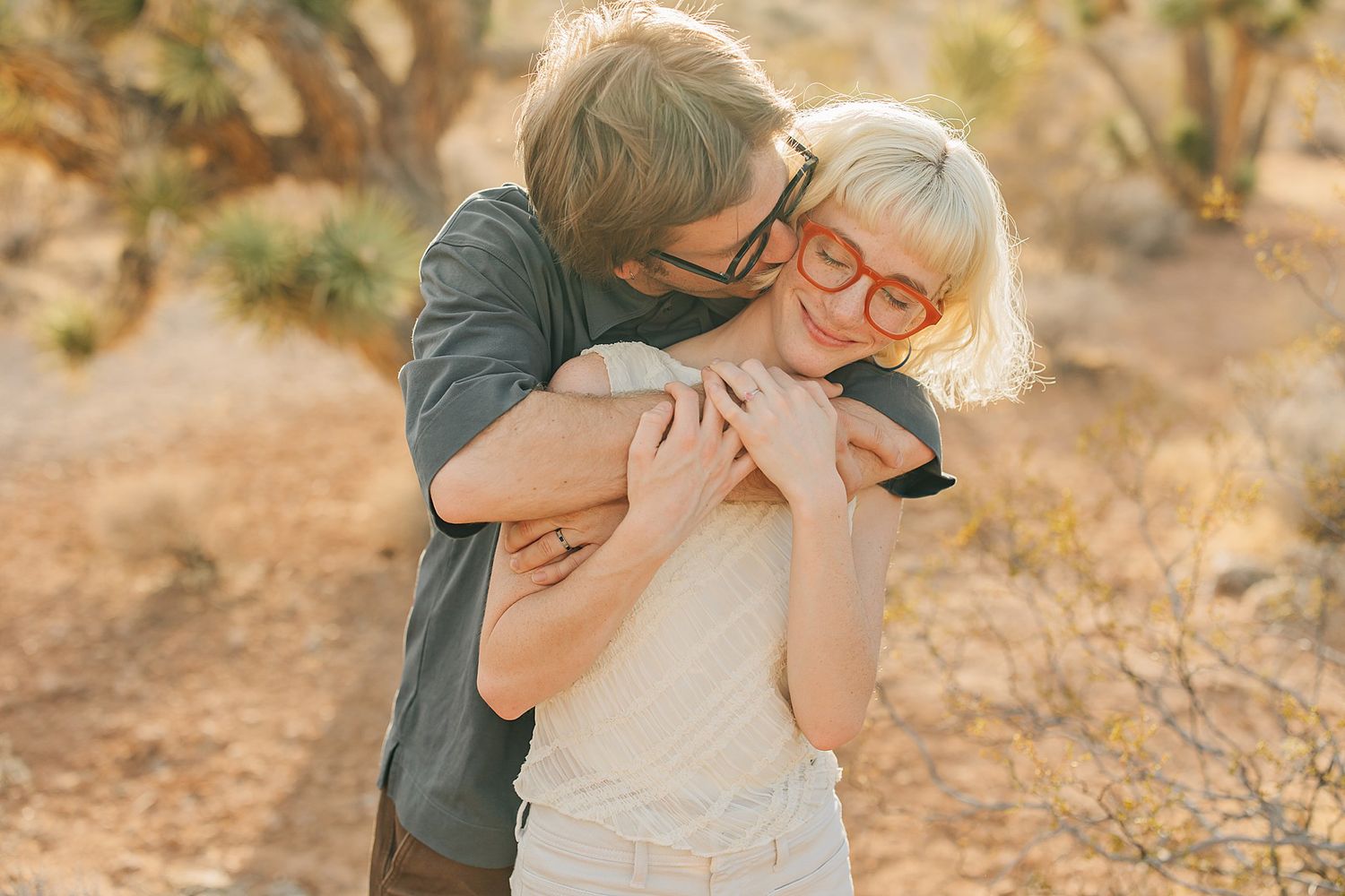 A couple embraces outdoors in a desert setting, woman wearing red glasses smiling joyfully.