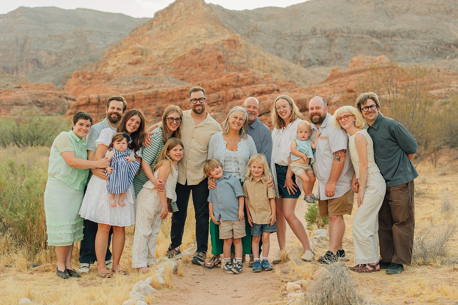 Large family group portrait smiling outdoors in desert landscape with red rock formations in background.