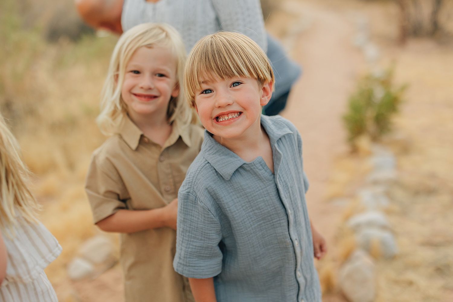 Two smiling blonde children in casual button-up shirts pose outdoors on a sunny dirt path.