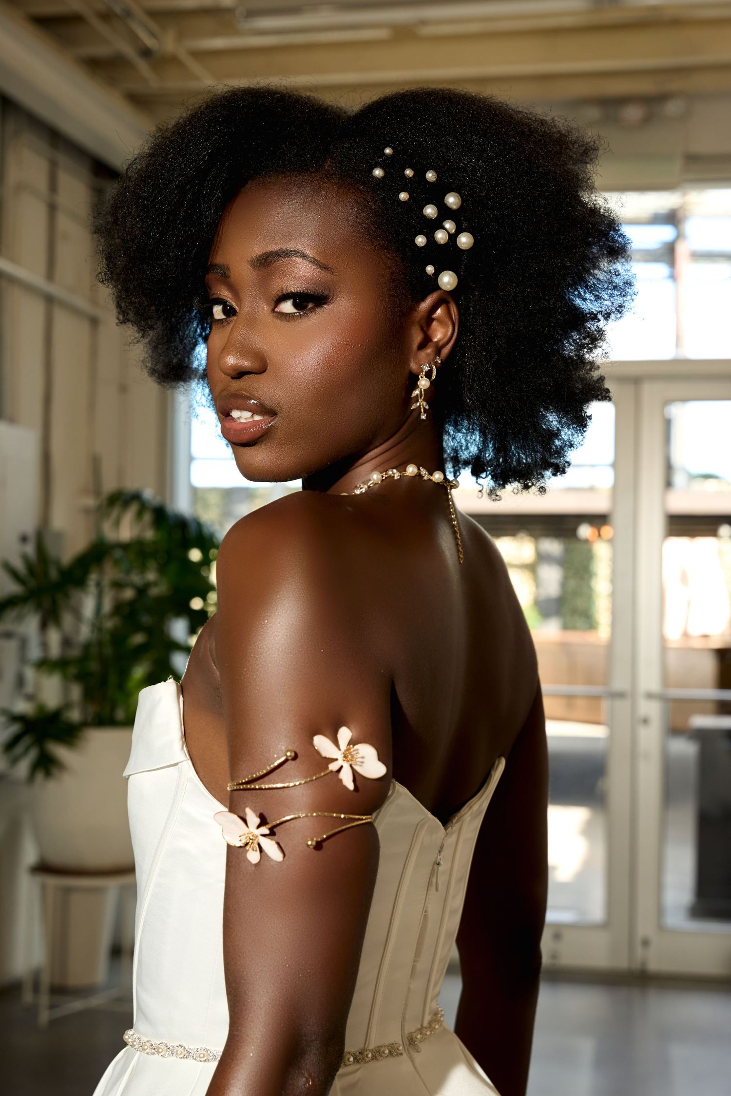 A young Black woman in a white strapless gown with floral arm jewelry and pearl hair accessories poses elegantly.