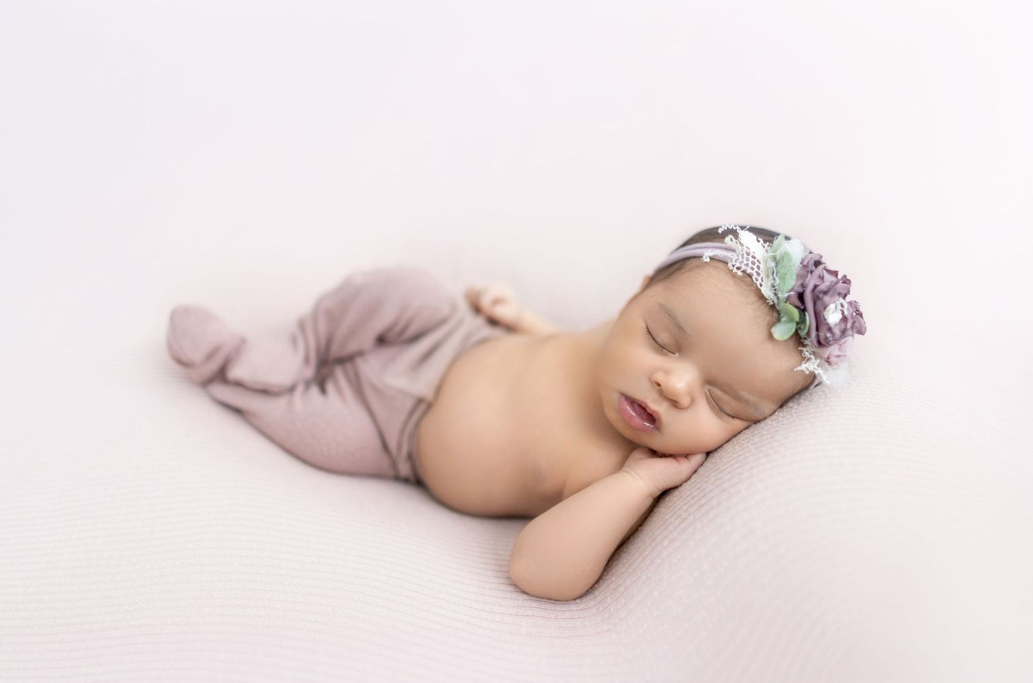 Sleeping newborn baby girl in mauve wrap and floral headband posed on white backdrop.