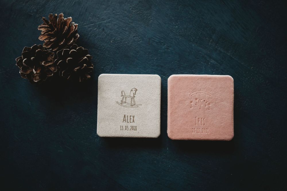 Two square photo boxes in beige and pink with pinecones on a dark surface.