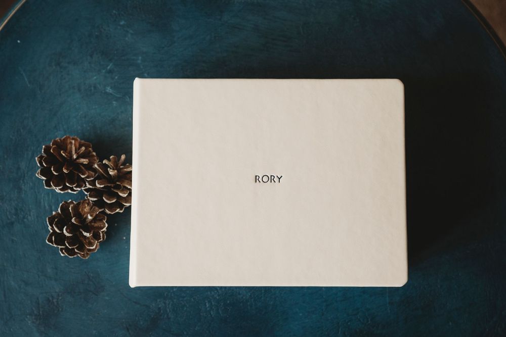 A white photo album labeled 'BORY' with pinecones on a blue surface.