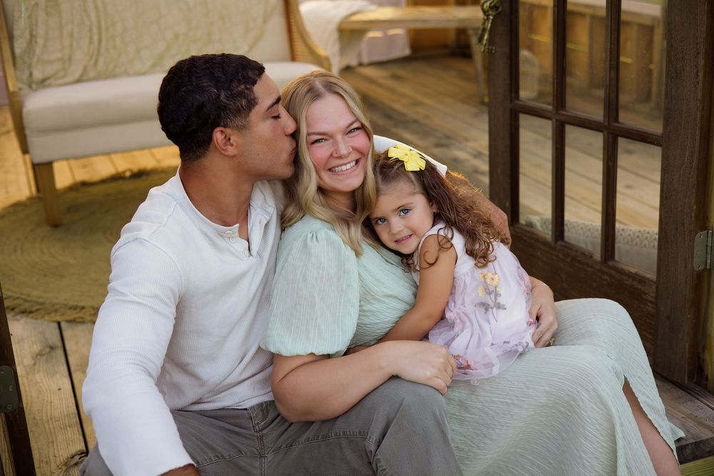 A happy family of three sitting together, with a man kissing a smiling woman's cheek while holding a young girl.