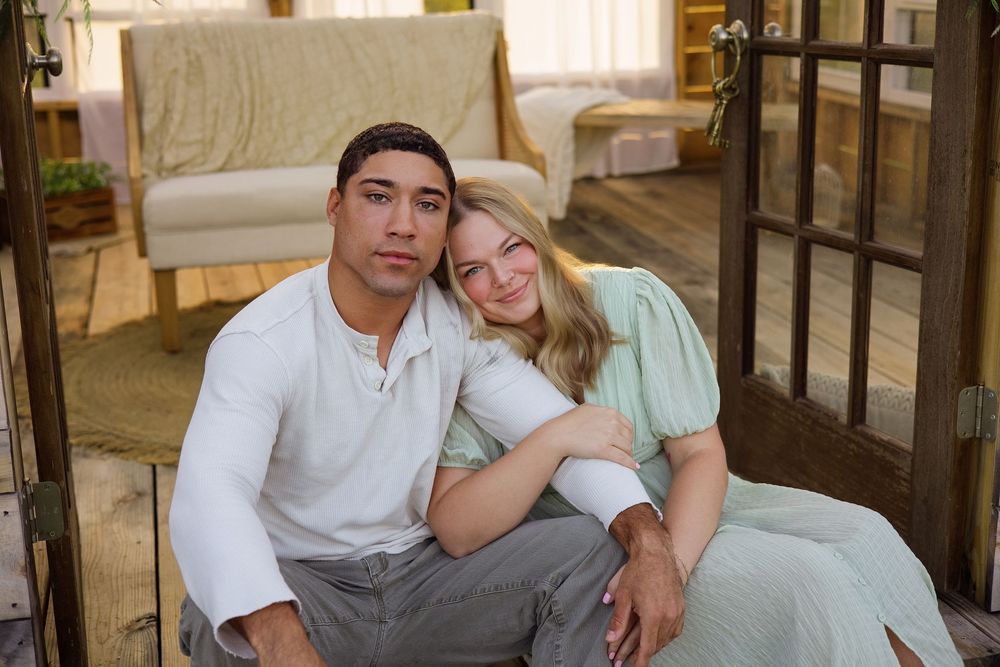 A young couple sitting together on a porch, smiling warmly, dressed in casual light-colored outfits.