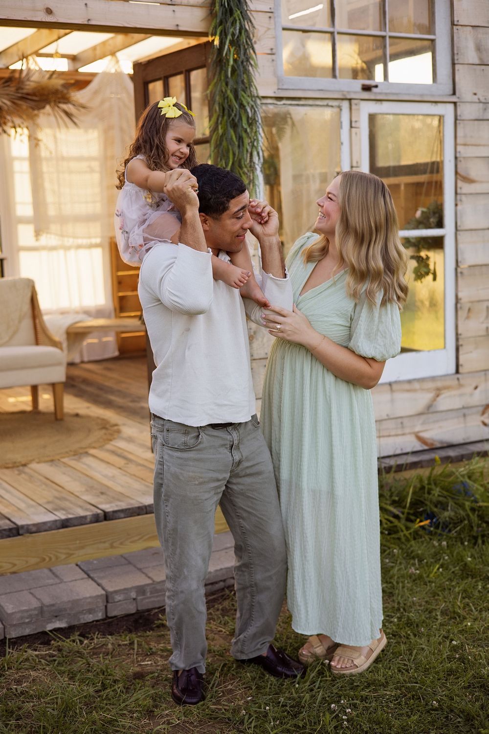 Happy family with young daughter on father's shoulders laughing outside their cozy wooden home.