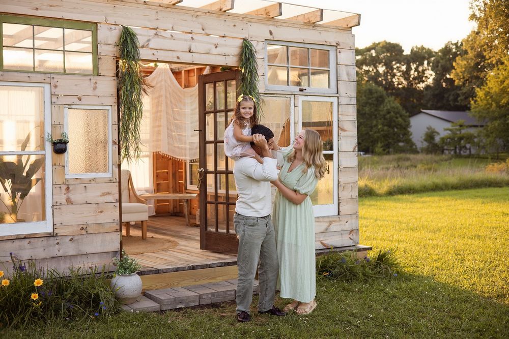 Happy family posing outside a rustic white farmhouse with greenery decor on a sunny day.