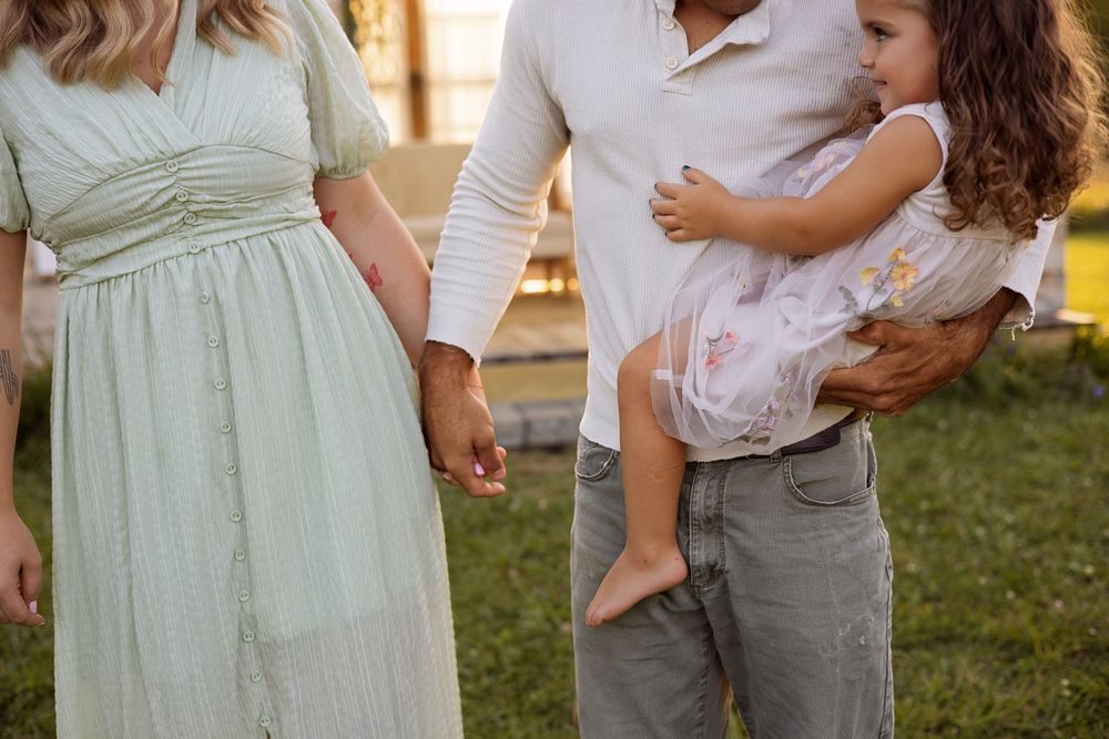 Pregnant woman in mint dress holds hands with man carrying toddler girl outdoors on green lawn.