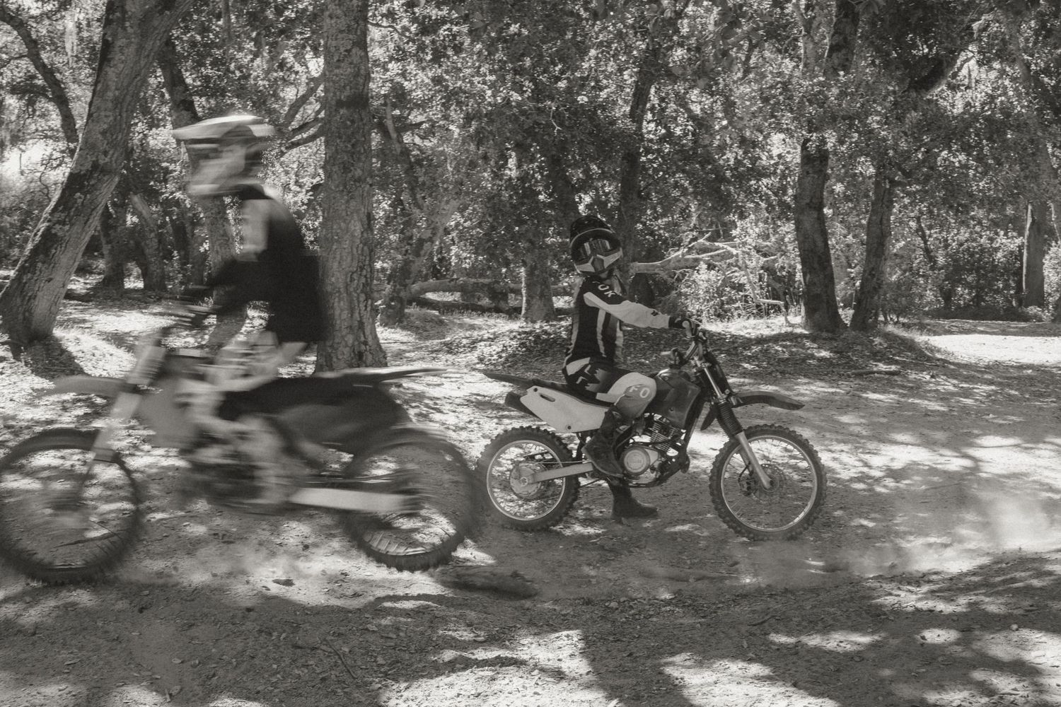 Two dirt bike riders race through a shaded forest trail in a vintage black and white action photo.