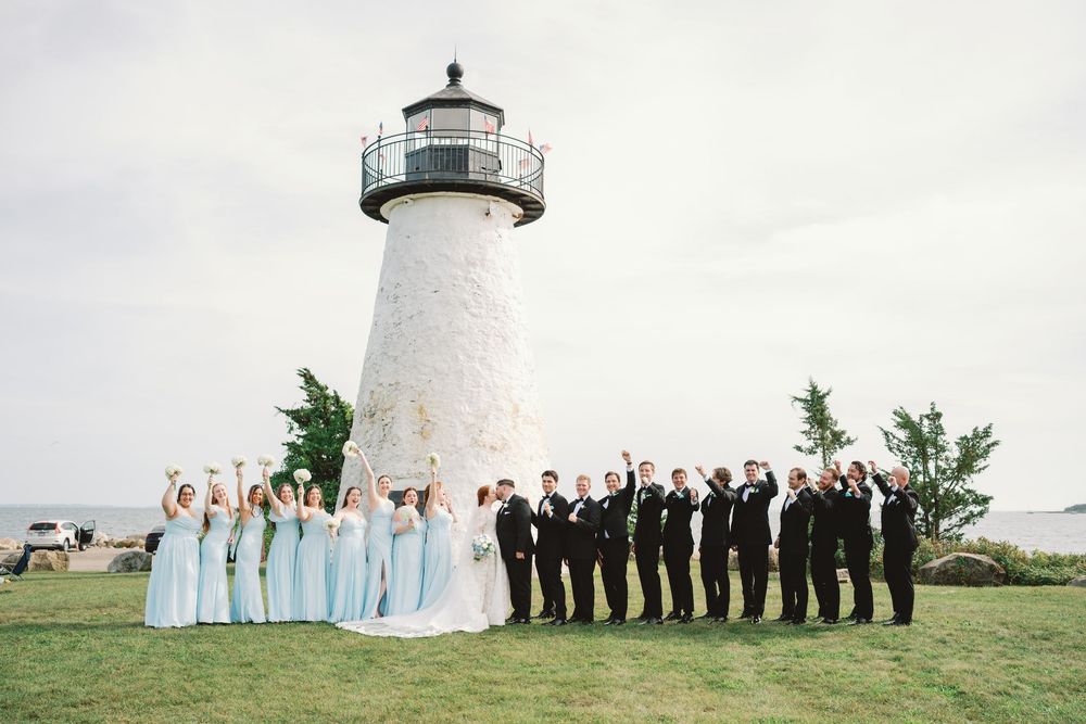 Wedding party celebrating in front of a white lighthouse, bridesmaids in mint gowns and groomsmen in black suits raising hands.