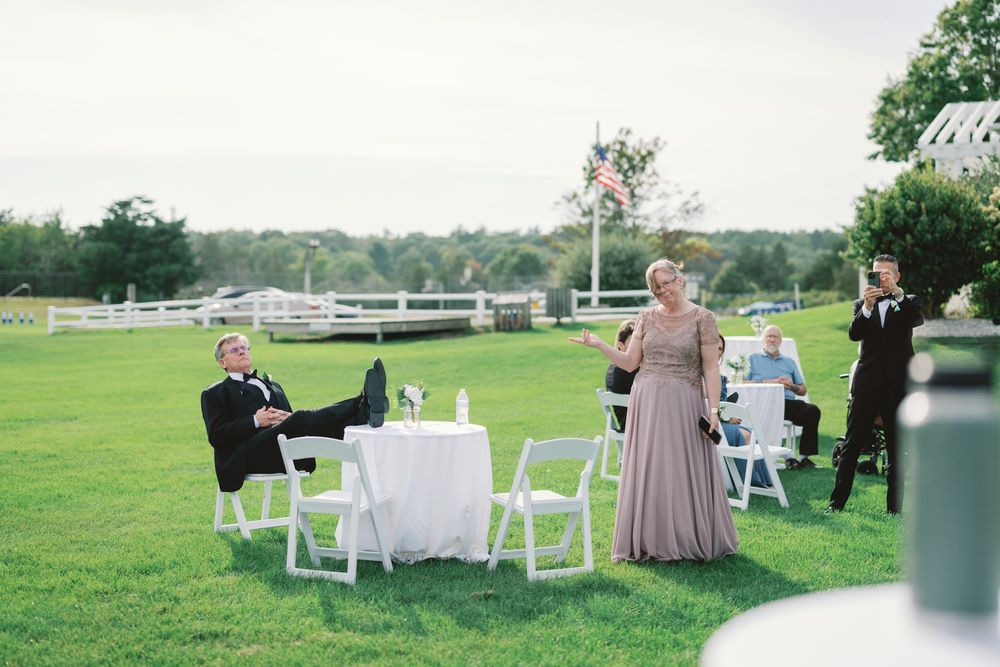 Outdoor wedding reception on green lawn with guests at white tables, woman in mauve gown speaking, man relaxing in chair.