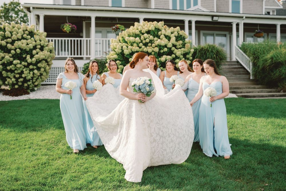 Bride in white lace gown twirls with bridesmaids in light blue dresses on green lawn before elegant house.