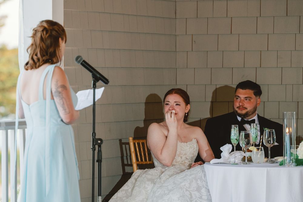 Bridesmaid gives a heartfelt speech at wedding reception while bride covers her mouth emotionally at the sweetheart table.