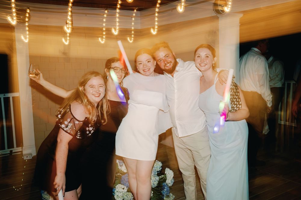 Bride in white dress celebrating with friends at wedding reception, holding glow sticks on dance floor.