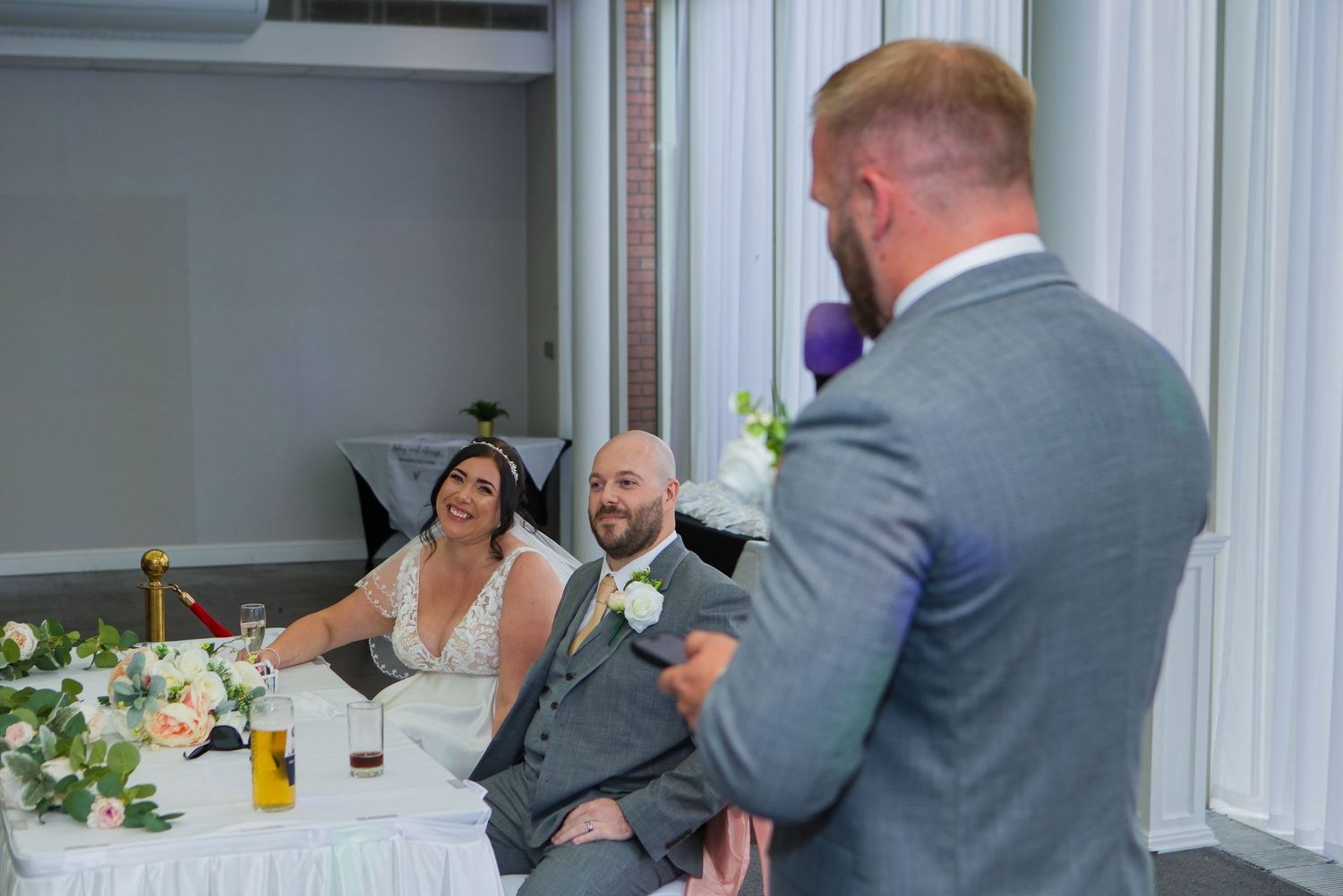 Bride and groom seated at wedding reception table smiling as best man delivers a heartfelt speech.