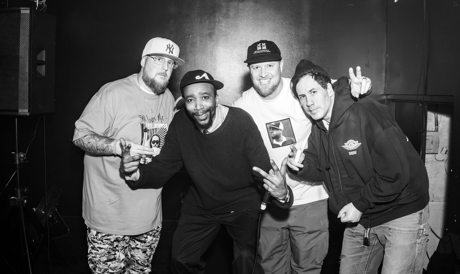 Four men posing together indoors, smiling and making hand gestures in a black and white photo.