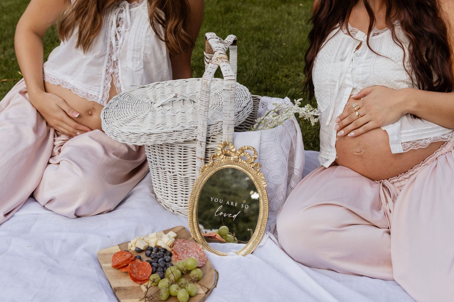Two women in pink and white dresses sit on grass during an outdoor maternity picnic photoshoot.