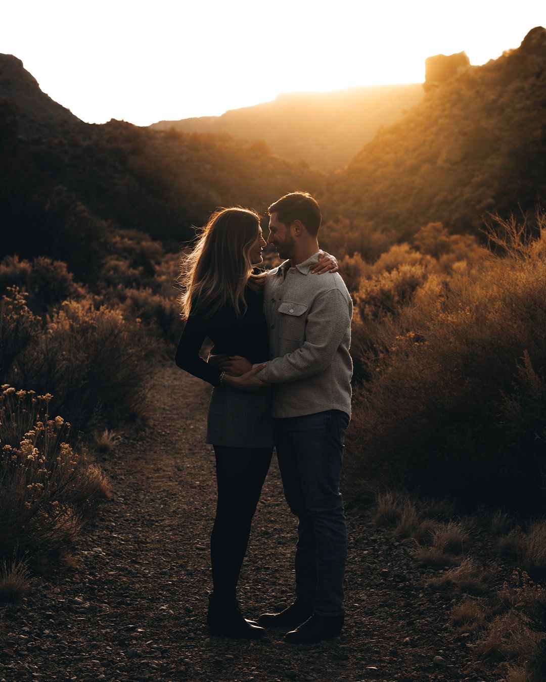 Couple embracing at golden sunset in a desert canyon landscape with warm backlight and rugged hills.