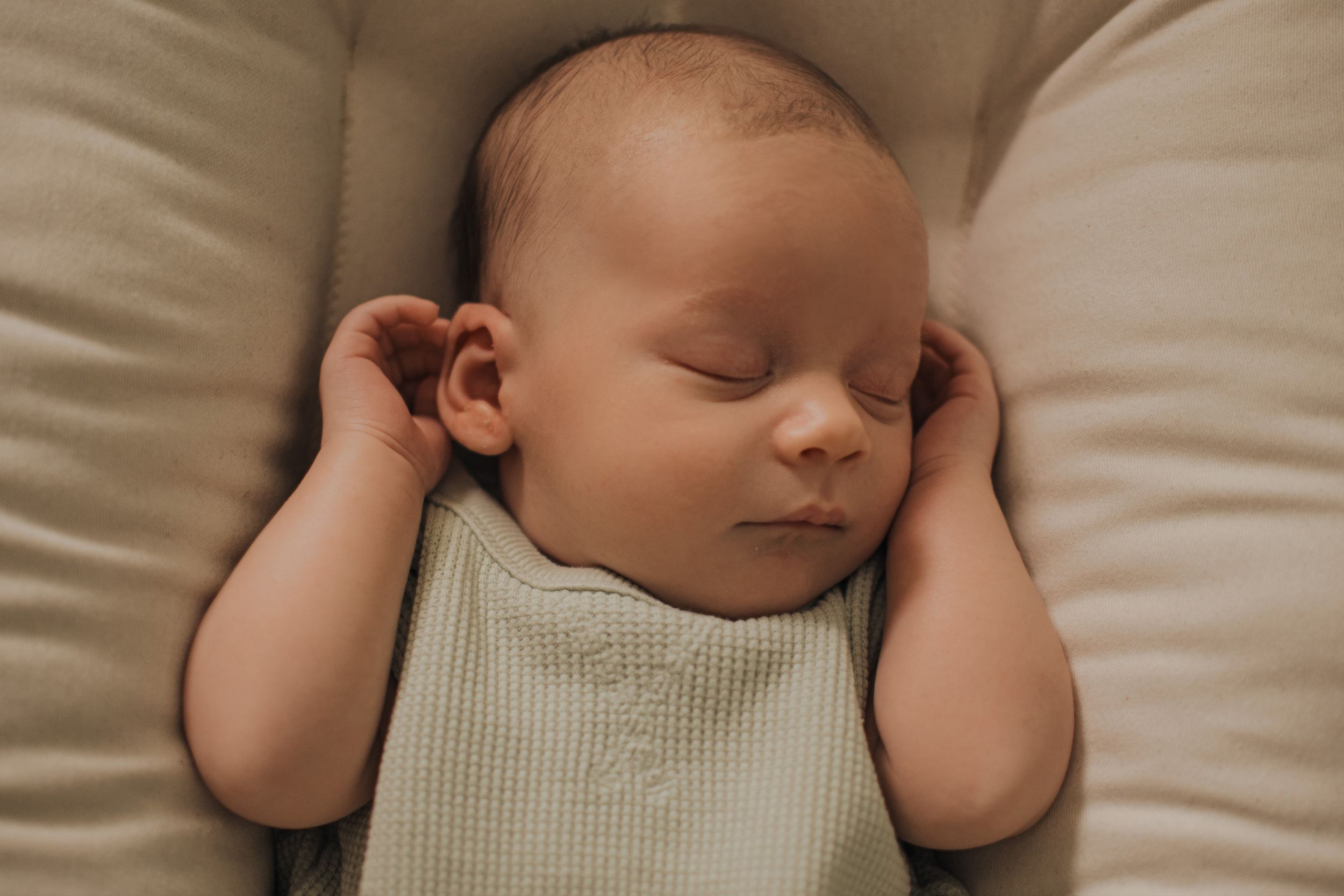 Peaceful newborn baby sleeping soundly in a soft beige nest, wearing a light green ribbed onesie with tiny hands raised.