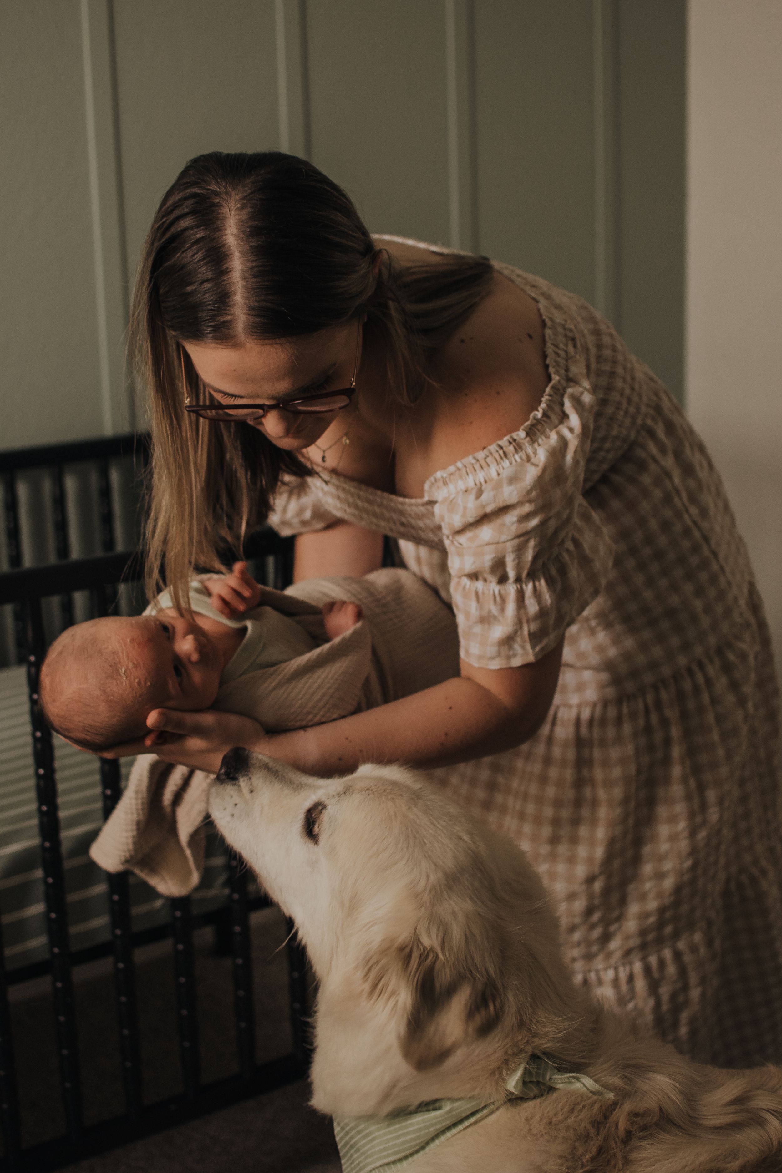 A young mother in a plaid dress kisses her newborn baby while a white dog sniffs the infant.