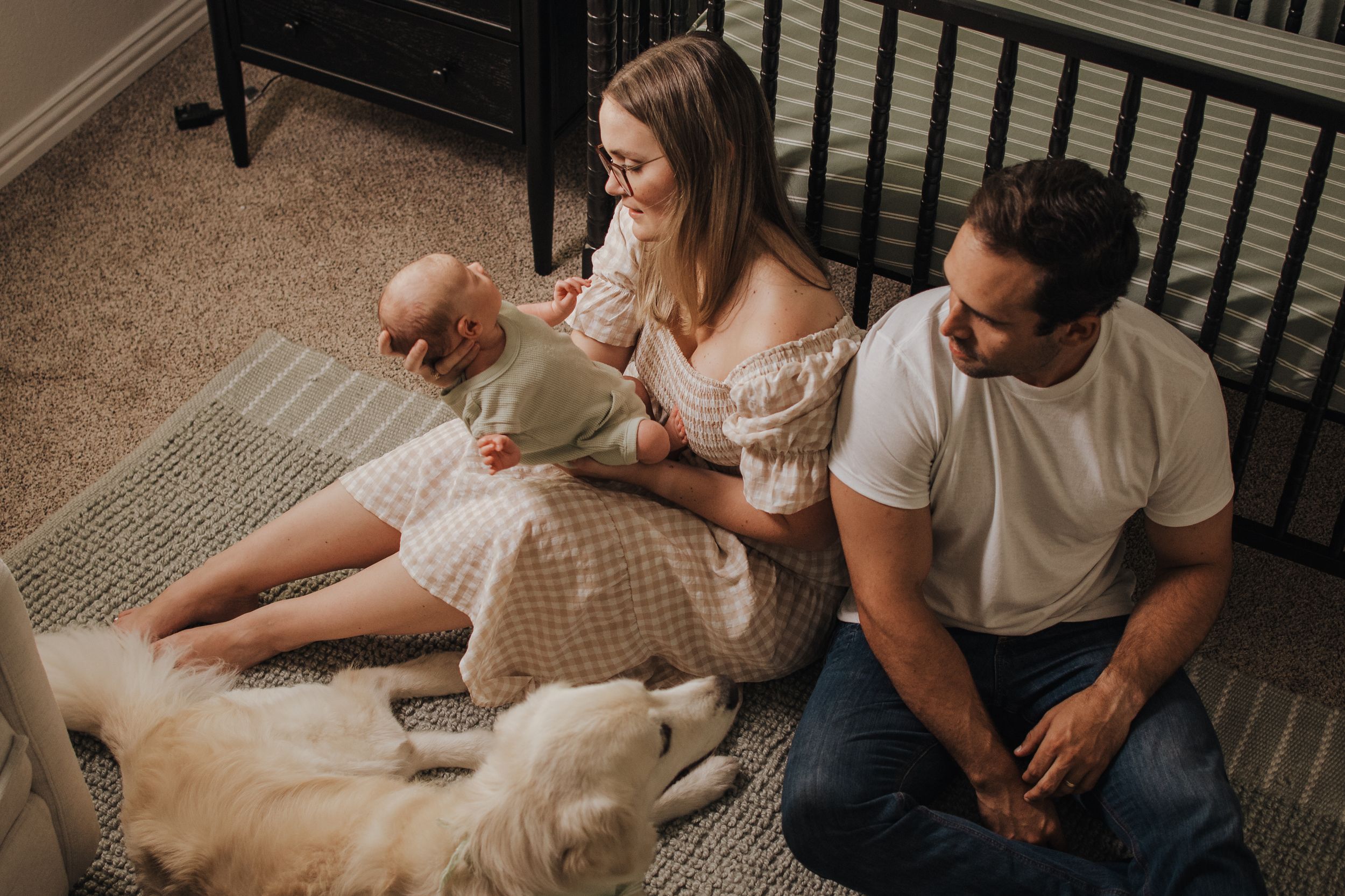 Young family with newborn baby and golden retriever dog sitting on nursery floor near wooden crib.