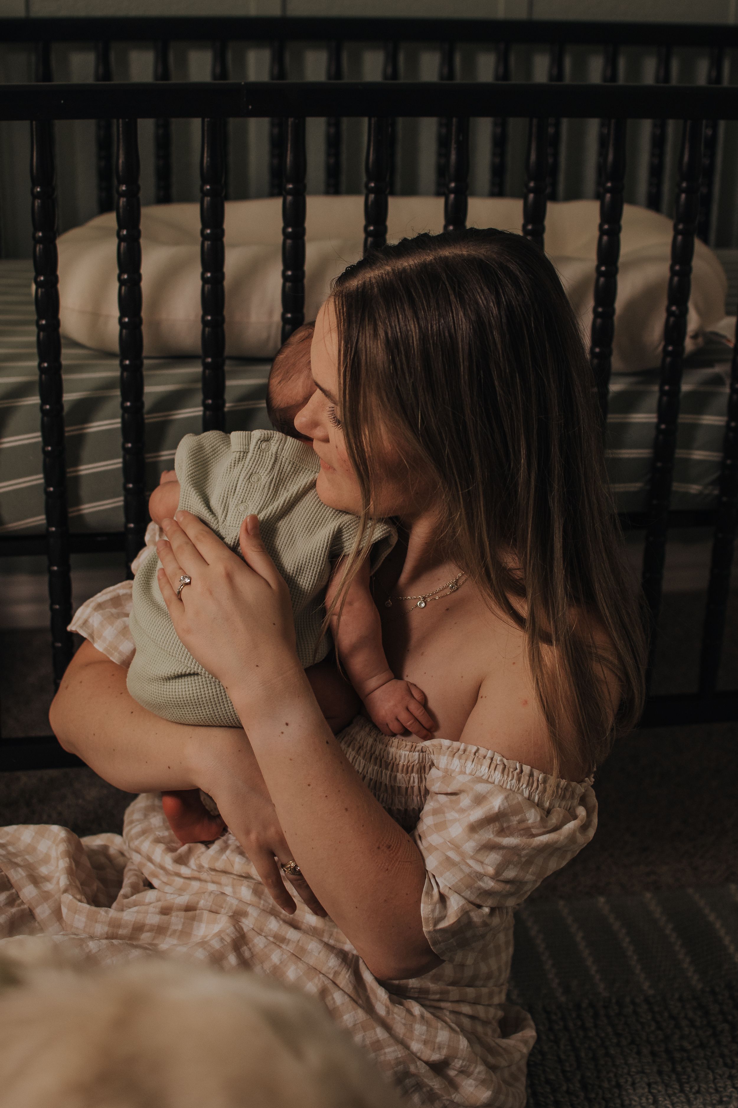 A mother tenderly holds her newborn baby close, sitting beside a dark wooden crib in a softly lit nursery.