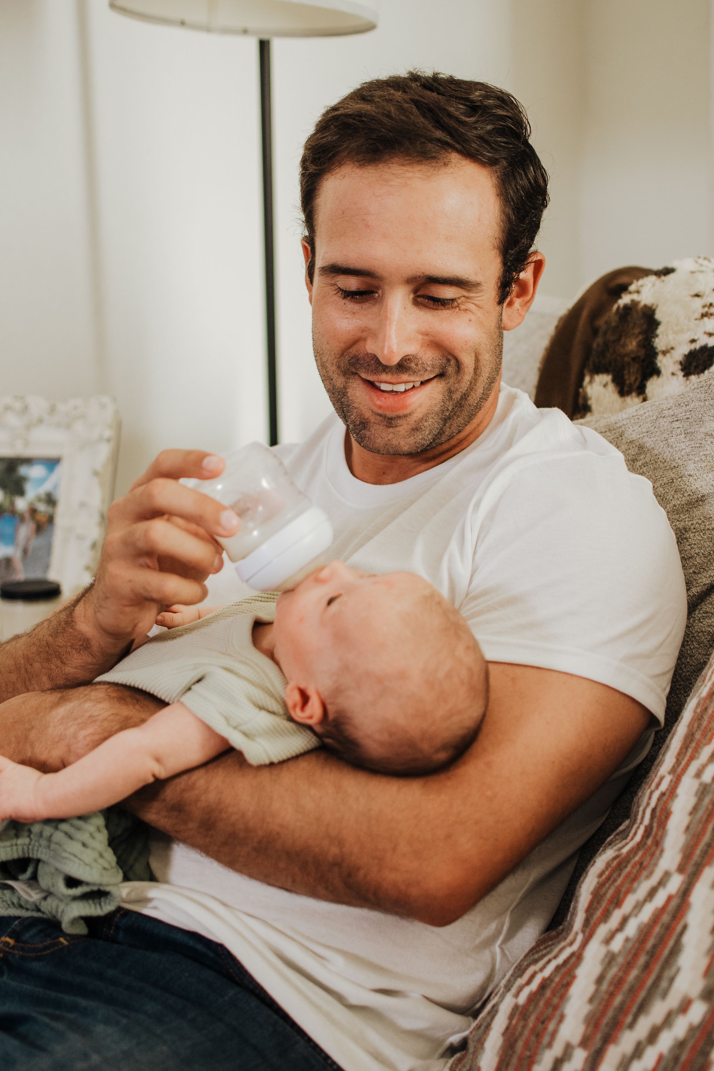 Smiling father bottle-feeding his newborn baby while sitting on a couch at home.