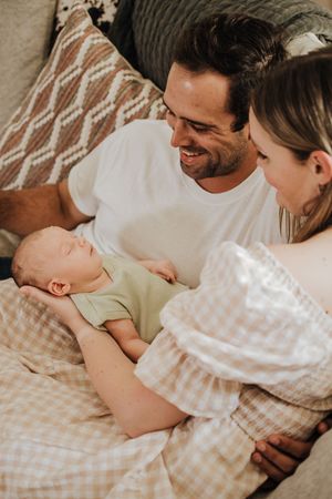 Happy parents smiling at their sleeping newborn baby lying on a gingham blanket at home.