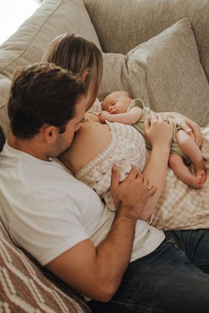 A couple sitting on a couch together, tenderly holding their sleeping newborn baby wrapped in a soft blanket.