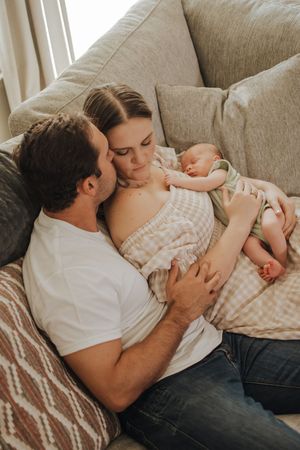 A couple cuddling on a gray sofa with their newborn baby, sharing a tender family moment in a cozy living room.