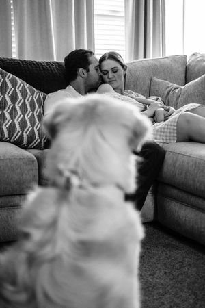 Black and white photo of couple cuddling on sofa while fluffy dog sits in foreground watching them.