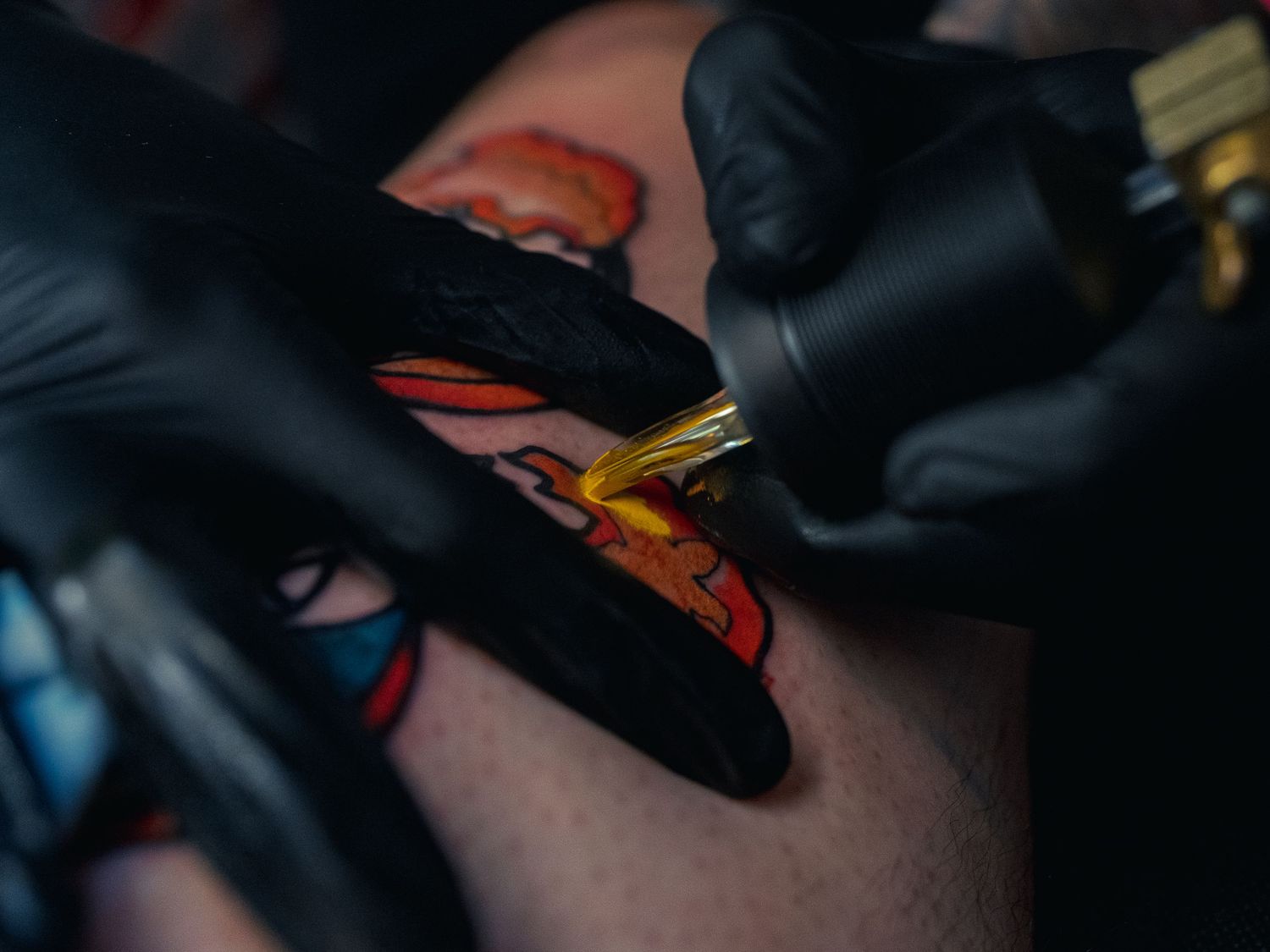 Black-gloved hands run a tattoo machine over skin, laying bold red, orange, yellow and blue ink into a fresh outlined design under low light