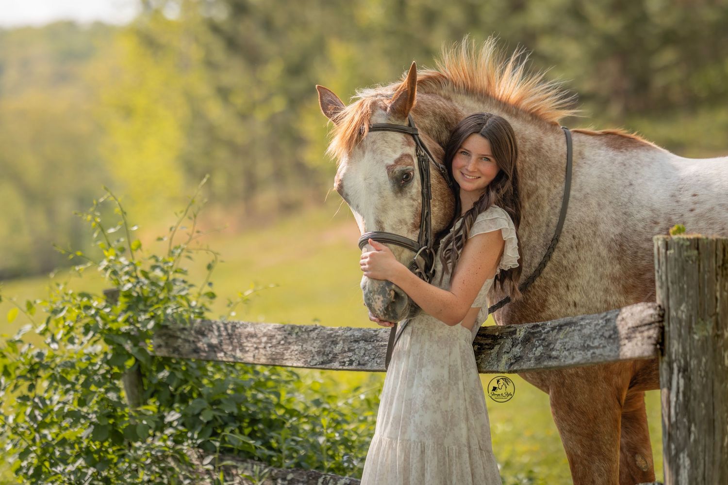 Young woman in white dress smiling beside a brown and white Appaloosa horse at a wooden fence outdoors.