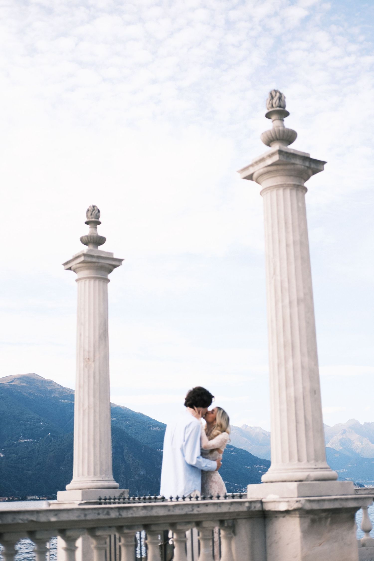 Couple embracing between elegant white columns on a terrace with mountain backdrop and cloudy sky.