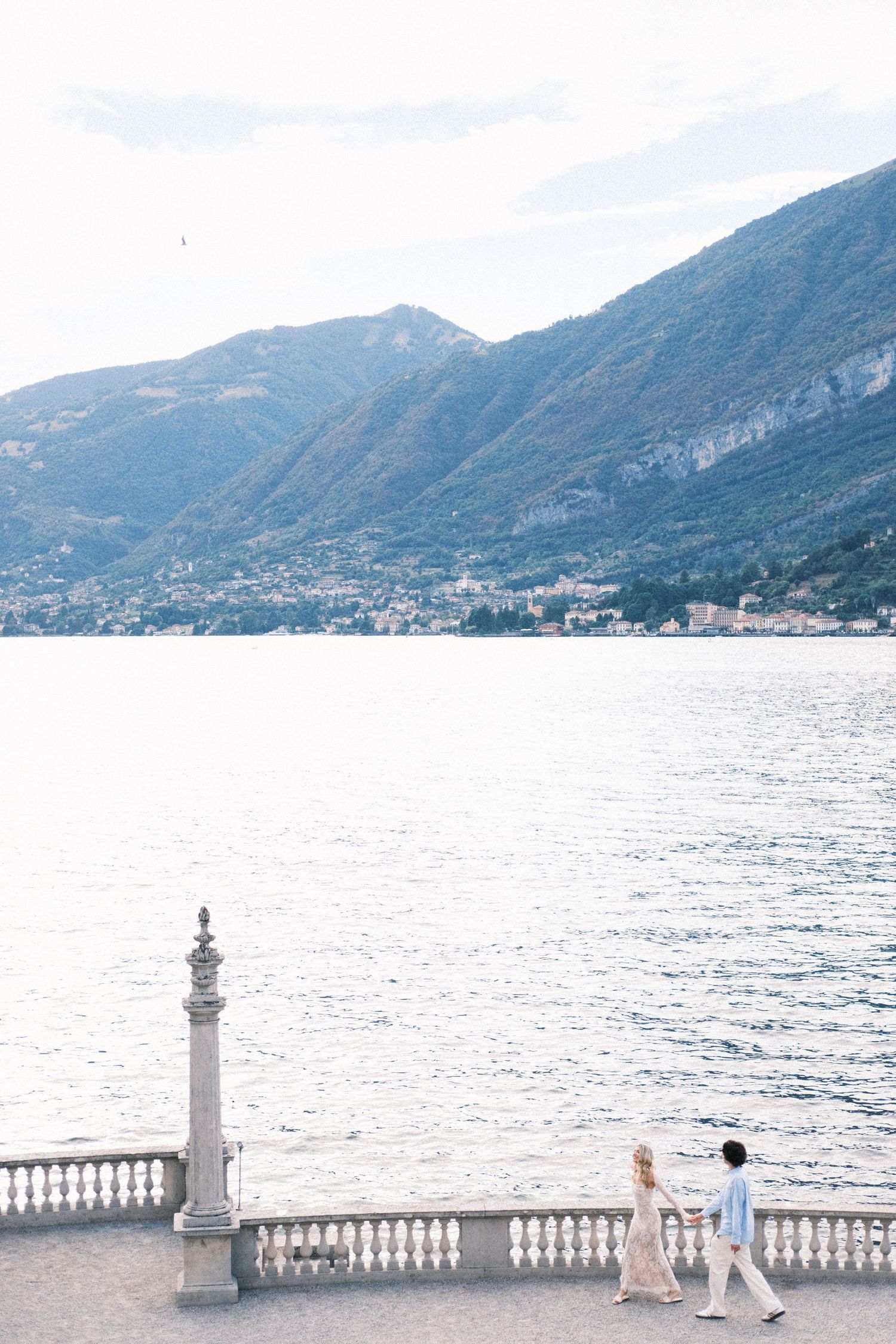 Couple walking hand in hand on a lakeside promenade with mountain views, Lake Como, Italy.