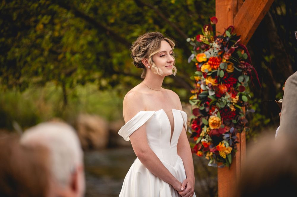 Bride in off-shoulder white gown stands at outdoor wedding altar with colorful floral wreath backdrop.