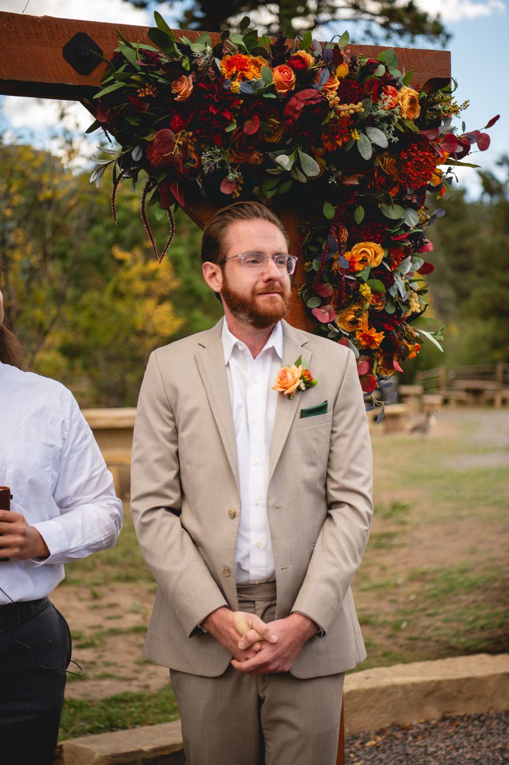 Groom in tan suit with orange boutonniere stands at outdoor fall wedding ceremony altar adorned with colorful floral arrangement.