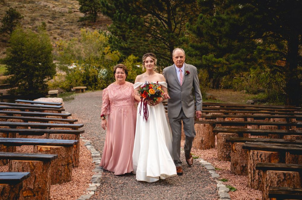 Bride walks down outdoor aisle with parents at rustic mountain wedding venue with stone benches and autumn foliage.