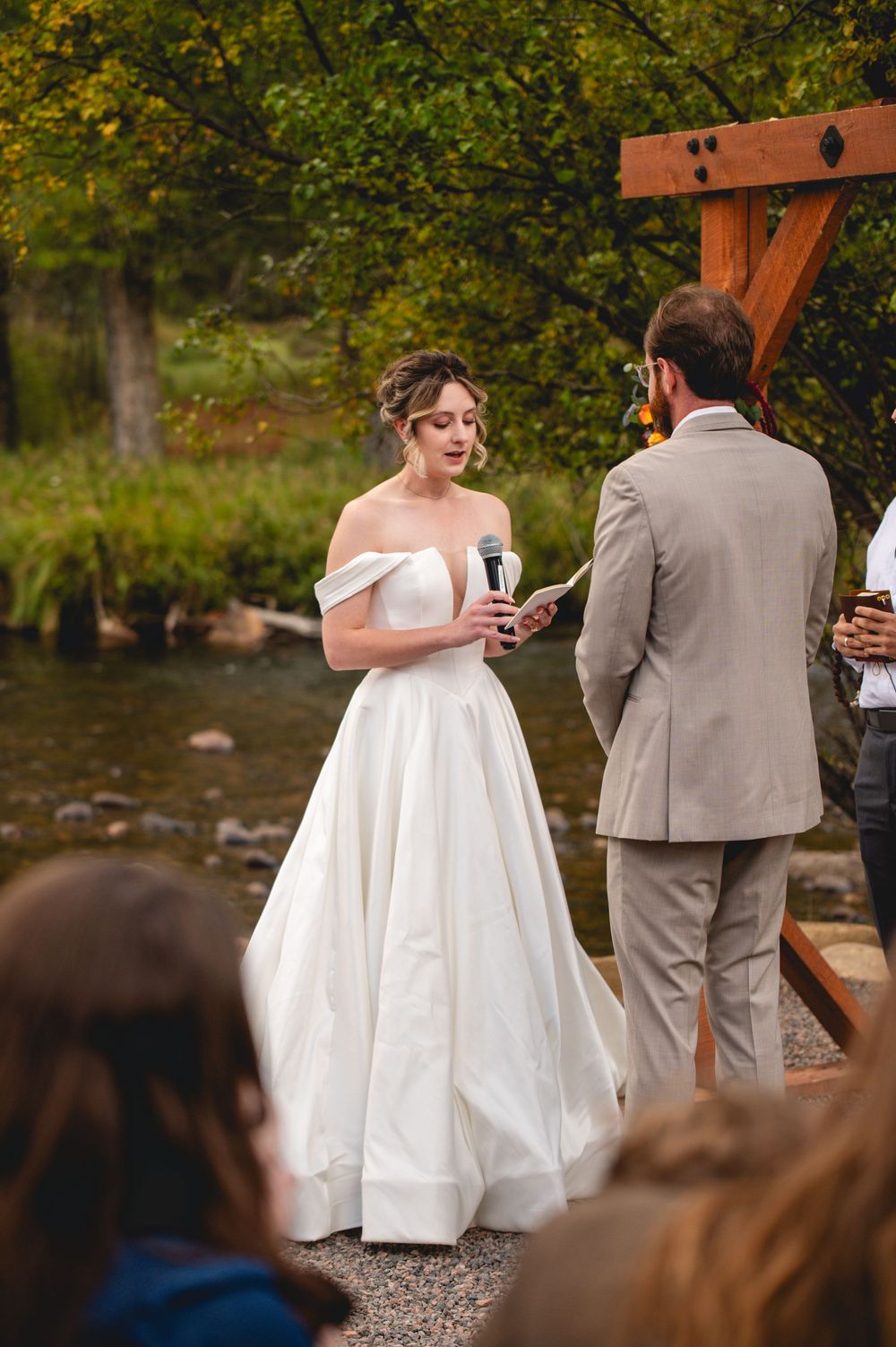 Bride in off-shoulder white gown reads vows outdoors by a creek, facing groom in tan suit at wooden arch.
